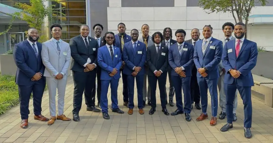 A group of 17 young African American men pose outdoors in front of a modern building with large glass windows and a white facade. They are dressed in formal business attire, wearing suits in shades of navy, black, and gray, paired with dress shirts and ties. Some of them have name tags pinned to their jackets. The group stands on a paved courtyard with greenery and a tree nearby, smiling confidently at the camera.