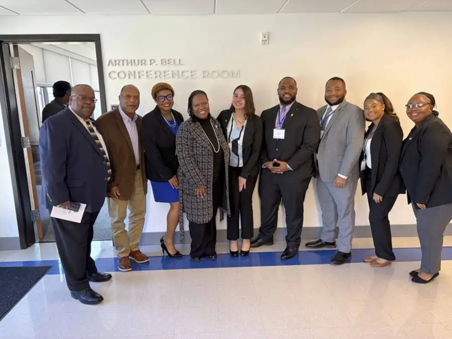 A group of nine professionally dressed individuals pose together in front of a wall with the sign Arthur P. Bell Conference Room. They are standing close together smiling at the camera. The group includes men and women of diverse backgrounds wearing business attire including suits, blazers, and dresses.
