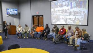 A group of people sit in a semicircle in a room with wooden doors and blue carpet, participating in a discussion. A woman in a black blazer and striped shirt stands on the left, holding papers. Eight individuals sit on chairs, one holding a microphone and speaking, wearing a brown vest over a plaid shirt. The others wear casual clothing, including a light-colored suit jacket, an orange shirt, a black beanie, a red shirt, and a brown leather jacket. A projection screen displays a collage of people, and a television screen shows a similar image. A wooden podium and a table with a yellow tablecloth are in the background.
