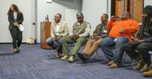 A group of people sit in a circle in a room with wooden doors and blue carpet, participating in a discussion. A woman in a black blazer and striped shirt stands on the left, holding papers. Five men sit on chairs, one holding a microphone and speaking, wearing an orange shirt. The others wear casual clothing, including a light-colored suit jacket, a green vest with a plaid shirt and cap, a brown jacket, and a black beanie. A wooden podium with a computer monitor is in the background.