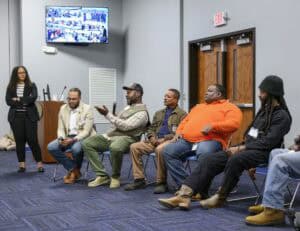 A group of people sit in a circle in a room with wooden doors and blue carpet, participating in a discussion. A woman in a black blazer and striped shirt stands on the left, holding a microphone. Five men sit on chairs, one holding a microphone and gesturing, wearing a plaid shirt and cap. The others wear casual clothing, including a green vest, an orange shirt, and a black beanie. A television screen on the wall displays a group of people.