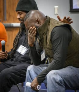 Two men sit in a room with wooden paneling, engaged in a discussion. The man on the left wears a black jacket, black beanie, and holds a microphone. The man on the right wears a green vest over a black shirt and jeans, holding a rolled-up document and gesturing with his hand.