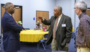 Three men stand in a room with wooden doors and blue carpet, engaged in conversation. The man on the left wears a blue suit jacket over a checkered shirt, the man in the center wears a gray suit jacket, white shirt, and striped tie, gesturing with his hand, and the man on the right wears a blue and yellow checkered shirt with khaki pants. In the background, people sit at a table with a yellow tablecloth.