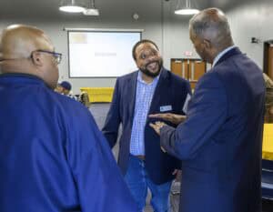 Three men stand in a room with wooden doors and a projection screen displaying "Welcome," engaged in conversation. The man on the left wears a blue jacket, the man in the center wears a blue blazer over a checkered shirt and jeans, and the man on the right wears a blue suit jacket over a checkered shirt. Yellow tablecloths cover tables in the background.