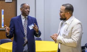 Two men stand in a room with a wooden door and a yellow tablecloth-covered table, engaged in conversation. The man on the left wears a blue suit, checkered shirt, and patterned tie, gesturing with his hands. The man on the right wears a light-colored suit jacket over a white shirt, holding his hands together.