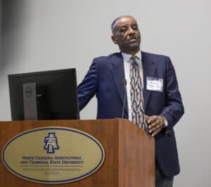 A man in a blue suit, checkered shirt, and patterned tie stands at a wooden podium with a microphone, speaking in a room. The podium has a sign that reads "North Carolina Agricultural and Technical State University Agriculture and Environmental Sciences." A computer monitor is on the podium.