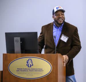A man in a brown blazer, blue checkered shirt, and cap stands at a wooden podium with a microphone, speaking in a room. The podium has a sign that reads "North Carolina Agricultural and Technical State University Agriculture and Environmental Sciences." A computer monitor is on the podium.
