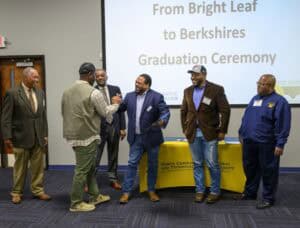Six men standing and conversing in front of a projector screen at a graduation ceremony for the "From Bright Leaf to Berkshires" program, hosted by North Carolina Agricultural and Technical State University.
