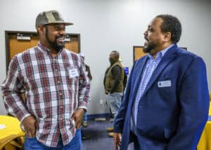 Two men stand in a room with blue carpet and wooden doors, engaged in conversation. The man on the left wears a plaid shirt, jeans, and a cap, with his hands in his pockets. The man on the right wears a blue suit jacket over a checkered shirt. Two other people are in the background near a table with a yellow tablecloth.