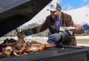 A man wearing black gloves and a brown blazer serves pulled pork from a large outdoor grill to someone holding a plate at a graduation event, with a white fence and blue sky in the background.