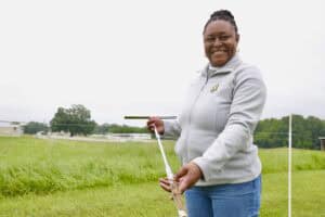 A woman smiling while holding a soil auger in a grassy field, wearing a light gray jacket with the N.C. A&T logo and blue jeans.