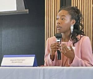 A young woman with dark, curly hair, wearing a pink blazer and large silver earrings, speaks into a microphone while seated at a panel discussion. She gestures with one hand as she speaks. In front of her, a nameplate reads "Beayonia Washington, Bennett College." The background features a modern conference room with wooden paneling.