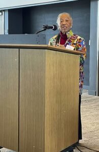 An older woman stands at a wooden podium speaking into a microphone, wearing a colorful patterned jacket and a name badge, inside a modern conference room.