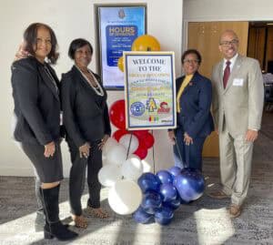 Four professionally dressed individuals stand together, smiling at the camera next to a welcome sign for the "HBCUs & Education: Social Justice Forum" event in the Deese Ballroom. The sign includes logos for North Carolina A&T State University, the NC Local Black College Fund, and Amazon. A cluster of red, white, and blue balloons is at the base of the sign. Behind them, a digital screen displays the hours of operation for the NC A&T Student Center.