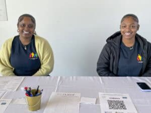 Two young women sit behind a registration table, smiling at the camera. They are wearing matching navy blue shirts with a colorful logo on the left side, one wearing a yellow cardigan and the other a black jacket. The table is covered with a white tablecloth and has sign-in sheets, a QR code agenda, pens in a small gold container, and a smartphone. A sign in the background is partially visible on the white wall.
