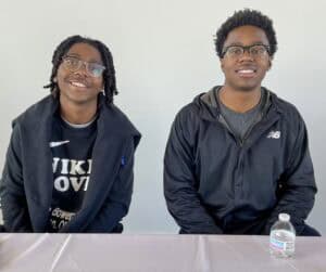 Two young men sit behind a table, smiling at the camera. The person on the left wears glasses, has short dreadlocks, and is dressed in a black Nike hoodie with a logo partially visible on the front. The person on the right wears glasses, has short curly hair, and is dressed in a black New Balance jacket over a gray shirt. A plastic water bottle is on the table in front of them. The background is a plain white wall.