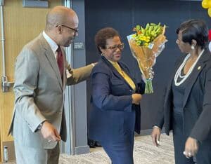 A woman in a navy blue suit and yellow blouse receives a bouquet of yellow and green flowers wrapped in brown paper. She is smiling while being congratulated by a man in a beige suit with glasses and a woman in a black suit with pearl accessories. The setting appears to be an indoor event space with a wooden door and dark wall panels in the background.