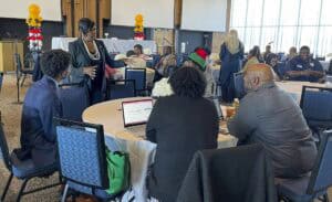 A professionally dressed woman wearing a black blazer and pearl necklace stands and speaks to a group seated around a round table covered with a white tablecloth. The table has a laptop, notebooks, and a floral centerpiece. The group includes individuals in business and casual attire, some taking notes or listening. In the background, other attendees are engaged in discussions at similar tables, with a stage, microphones, and balloon decorations visible in the conference room.