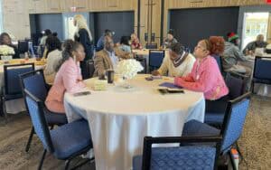 A group of four people sits around a round table covered with a white tablecloth in a conference room, engaged in discussion. The table has a floral centerpiece and various notebooks, pens, and a coffee cup. One woman in a pink blazer is speaking, while another in a pink sweater listens attentively. A man in a beige blazer and a woman in a cream sweater take notes. In the background, other attendees are seated at similar tables, conversing and working.