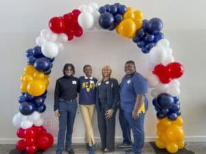 Four young adults stand side by side, smiling at the camera under a colorful balloon arch made of red, white, blue, and yellow balloons. Two individuals wear clothing featuring the North Carolina A&T State University logo, while the others are dressed in professional attire. The background is a plain white wall.