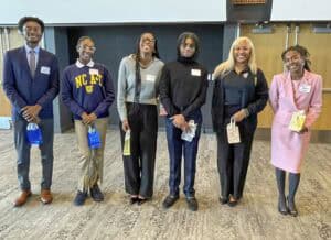 Six individuals stand side by side in a conference room, smiling at the camera while holding small gift bags. They are dressed in a mix of business and casual attire, with one person wearing an "NC A&T" sweatshirt and another in a pink blazer dress. The group consists of young adults and a professional with blonde hair. The background features wooden doors and a dark-paneled wall.