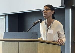 A young woman wearing glasses and a cream-colored sweater speaks at a wooden podium with a microphone in a modern conference room.