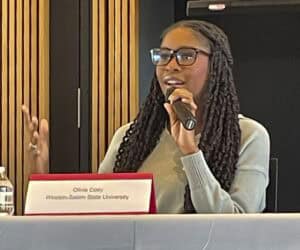 A young woman with long braided hair and glasses speaks into a microphone while seated at a panel discussion. She is wearing a light gray sweater and gestures with one hand as she speaks. A nameplate in front of her reads "Olivia Cody, Winston-Salem State University." The background features a modern conference room with wooden paneling.