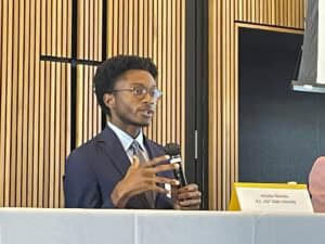 A young man with short curly hair, glasses, and a neatly trimmed beard speaks into a microphone while seated at a panel discussion. He is wearing a navy blue suit, a light blue dress shirt, and a gray tie. He gestures with one hand as he speaks. A nameplate in front of him reads "Winston Wardlaw, N.C. A&T State University." The background features a modern conference room with wooden paneling.