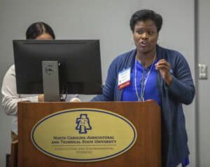 A woman wearing a blue blazer and a name tag labeled "Speaker" is standing behind a wooden podium with the North Carolina Agricultural and Technical State University logo. She is speaking into a microphone and gesturing with her right hand. A second person, partially obscured, is standing behind her working on a computer monitor.