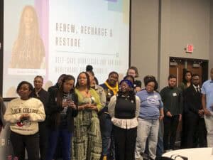 A diverse group of people stands in a conference room in front of a large projector screen displaying a presentation titled "Renew, Recharge & Restore: Self-Care Strategies for Aspiring Healthcare Professionals" by Ryanne Peterson. The attendees, dressed in casual and semi-formal attire, appear engaged, with some looking at their phones, clasping their hands, or listening attentively. The setting includes a neutral-colored wall, double doors, and a visible exit sign in the background.