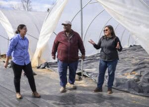 Three people stand and converse inside a greenhouse with translucent walls. The floor is lined with black material. One person gestures with their hands, and the others listen attentively. They are casually dressed and appear engaged.