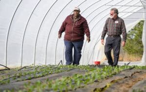 Two men walking in a greenhouse filled with young plants. The man on the left wears a maroon jacket and cap, while the man on the right wears a plaid shirt and sunglasses. Rows of small plants are visible under a curved, translucent roof.