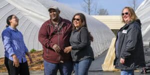 Four people are laughing and standing in front of a partially constructed greenhouse. Theyre outdoors, wearing casual clothing, and appear to be enjoying each others company. The background shows plastic greenhouse coverings.