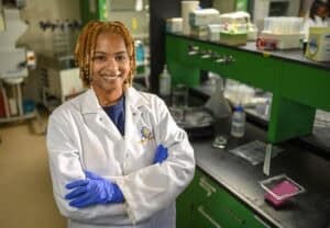 A woman wearing a white lab coat and blue gloves smiles confidently with arms crossed in a science laboratory with green cabinets and lab equipment on the counters.
