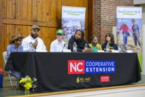 A panel of six individuals sits behind a table with an NC Cooperative Extension banner at a Small Farms Week event. One man speaks into a microphone while others listen. The panel includes youth and adults of diverse backgrounds. Behind them are banners promoting Small Farms Week, with images of farming activities. A vase of yellow flowers sits in front of the table.