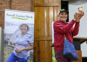 A person in a red hoodie and cap is smiling and holding up a small decorative object. They are standing next to a Small Farms Week banner featuring a photo of another person with plants.
