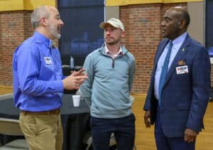 Three men are standing and talking in a room with brick walls. The man on the left is wearing a blue shirt, the middle man a light blue sweater and cap, and the man on the right a blue suit. They appear to be engaged in conversation.