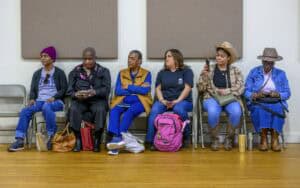 Six people sit on chairs against a wall in a room. They are wearing casual clothing, including hats and jackets. Some are looking at their phones, while others have their hands in their laps. Bags are placed on the floor in front of them.