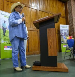A person in a cowboy hat and denim outfit speaks into a microphone at a wooden podium. Behind them are banners about Small Farms Week. The room has wooden paneling and some chairs.