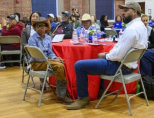People seated at round tables during an indoor event, focusing on a child in a cowboy hat and a man next to him. They are both wearing casual attire. Other attendees are visible in the background, and the tables are decorated with red cloths and flowers.