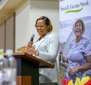 A smiling speaker holds a microphone at a podium during an event. A banner behind her reads Small Farms Week and features an image of a woman holding vegetables, underlining an agricultural theme.