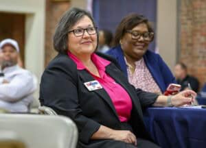 Two women sitting at a table during an event. The woman in the foreground wears a bright pink shirt and a black blazer with a name tag, holding a pen. Another woman sits beside her, smiling and using a phone. People are in the background.