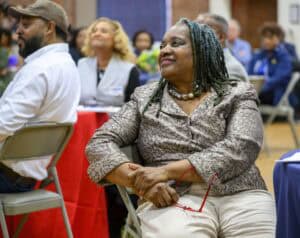 A woman with braided hair, wearing a patterned blouse and holding red glasses, sits attentively at an event. She smiles while looking towards the stage. Other attendees are seated around her in a hall with red and blue tablecloths.