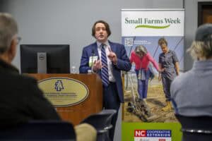 A man in a suit speaks at a podium with the North Carolina A&T logo. A Small Farms Week banner is displayed behind him. Audience members sit facing him. A screen is partially visible to the left.