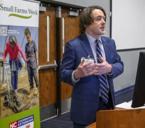 A man in a blue suit and striped tie speaks at a podium. Behind him, a banner reads Small Farms Week and shows two people farming. A logo for NC Cooperative Extension is visible at the bottom of the banner.