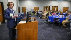 A man in a suit speaks at a podium in a conference room. Attendees sit at tables with blue tablecloths, listening attentively. The room has neutral walls, and doors are visible in the background.