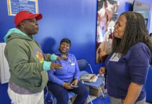 Three women engaged in conversation indoors. One in a red cap and green hoodie gestures while speaking. Another, in a blue shirt, sits and smiles. The third, with long hair, stands and listens attentively. A blue wall and poster are in the background.