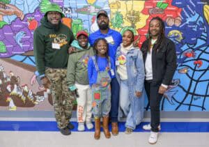 A group of six people stands smiling in front of a colorful mural. They are casually dressed and appear diverse in age and style. The mural depicts various animals and geographical shapes, adding a vibrant backdrop to the scene.