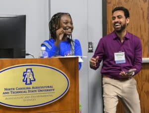 A young girl smiles and speaks into a microphone at a podium with a North Carolina Agricultural and Technical State University logo. A man beside her laughs and applauds. Both appear to be enjoying the moment.