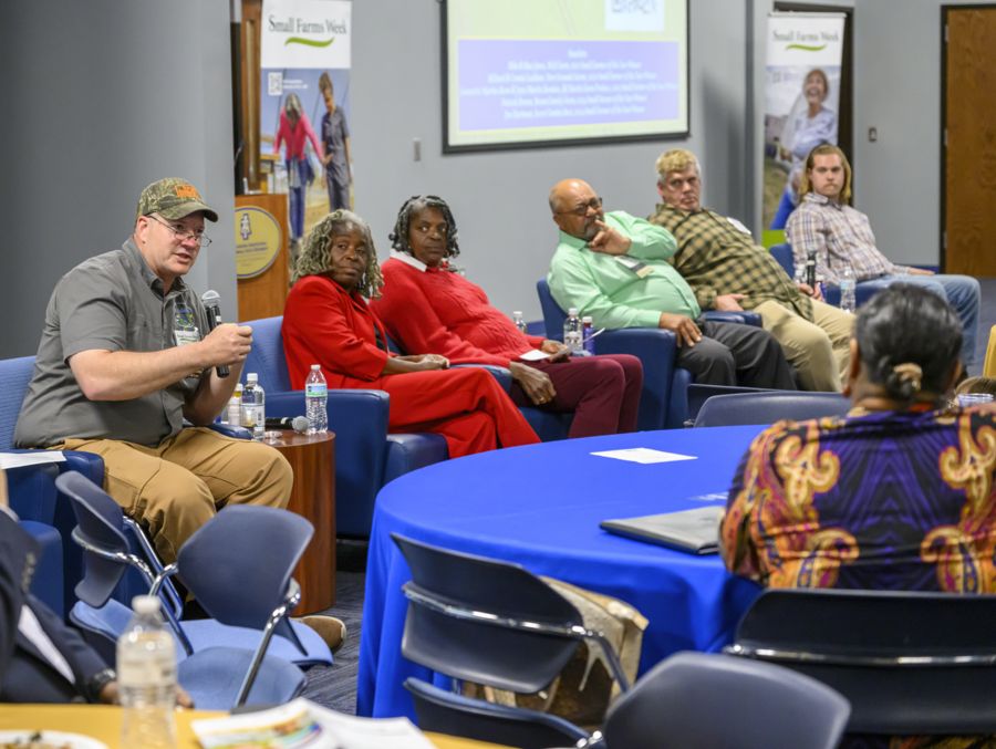 A diverse group of people sit on a panel in a conference room, engaging in a discussion. A man in a cap speaks into a microphone, while others listen. A presentation screen and banner are visible in the background. The room has blue-covered tables.